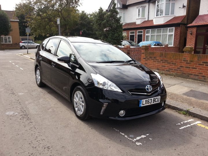 Toyota Prius Plus MPV - anyone got one? - Page 1 - Jap Chat - PistonHeads - The image shows a black Toyota Corolla LX6 driven by a person. It is parked on the street beside a curb, adjacent to a sidewalk and a residential house with a brick wall. The vehicle has a white license plate, and there are trees in the background. The Toyota logo is visible on the front grille. The car appears to be in a residential area.