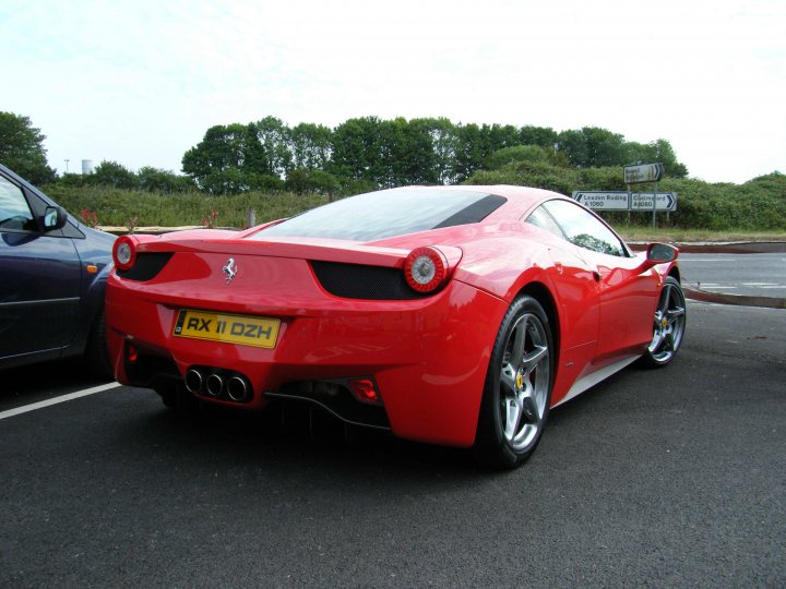 Breakfast Meeting, The Hare, Roxwell 4th June - Page 1 - Kent & Essex - PistonHeads - In the image, a red Ferrari sports car is parked in a gravel parking lot. The car is facing the left side of the image and is distinguished by its license plate, which reads "T60HZH". The license plate and the car's design suggest it's a European model. There are other cars parked in the distance, with a night sky visible in the top left corner. The text "W60 WSK0 W6T MCS9" indicates a location or destination, possibly suggesting the image was taken near a motorway exit. The overall scene suggests a road trip or a parking area at a destination point.