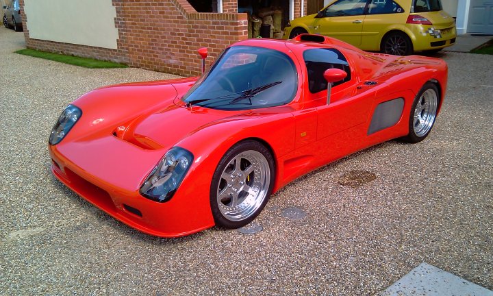 Garage Finally Pistonheads Ultimawell Birth - The image showcases a vibrant red sports car parked on a gravel surface. The car's distinctive design features a pop-up windshield and targa top, offering an open-air driving experience. The vehicle's elegant curves and advanced aerodynamics hint at its high-performance capabilities. Parked in the background is another car, which contrasts with the red sports car in terms of color and body style.