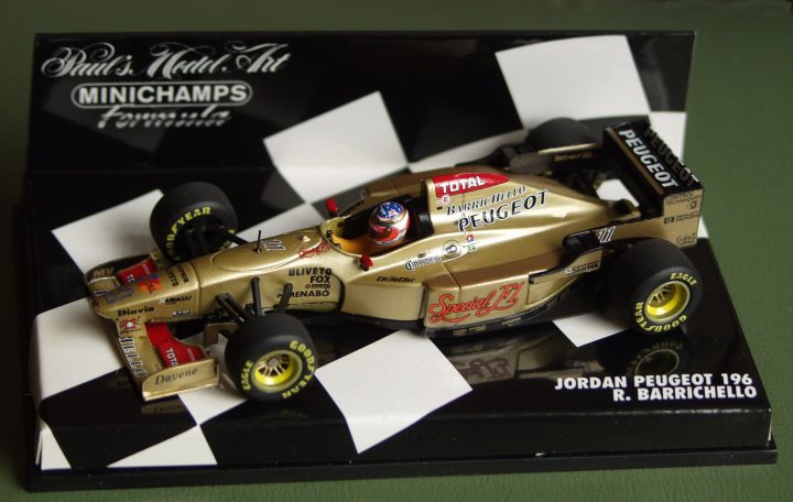 Pistonheads Collection - The image presents a scale model of a race car on a checkered flag background. The car, modeled as an F1 racer, is predominantly gold with black accents. It features sponsorship logos including "Total" and "Cooper Tyres." The driver figure in the cockpit is wearing a racing suit with the name "Jordan" and the number 196. The model is displayed on a black, checkered flag base, designed to mimic the appearance of a racing podium. The overall image suggests a collector's item or a souvenir for a racing fan.