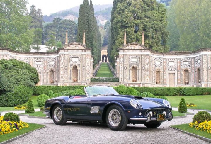 A car parked in front of a building - Pistonheads - This image portrays an historic, albeit partly ruined, structure with classical architecture, set against a backdrop of verdant trees and imposing mountains. The central focus is an antique, convertible sports car, which is parked with its top down, on a gravel road. The entrance to the building features a grand, tree-lined driveway leading up to the structure, which is adorned with various decorative elements and statues. The image evokes a sense of nostalgia and the romance of grand estates in a serene landscape.