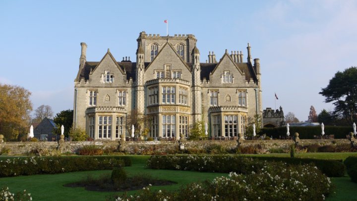 Btap Pistonheads Polaroids - The image shows a large, historic castle-like building, which is likely a hotel or inn. The structure is multi-winged with numerous windows and dormer windows on the roofs. It is surrounded by well-manicured gardens with various pathways leading to and around the building. Within the foreground, there are well-defined hedges and cultivated flowers. The sky is clear and blue, indicating a sunny day.