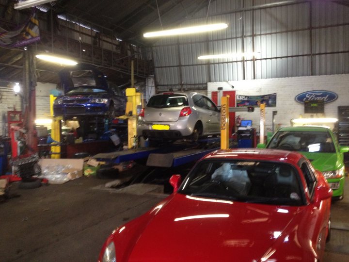 Wheel Alignment Recommendation - Page 1 - South West - PistonHeads - The image shows a busy car repair shop with a variety of vehicles and tools. In the middle of the frame is a silver hatchback car on a lift, being serviced along with other cars visible in the background. To the right, there's a green car parked off to the side next to a blue truck. The parking lot of the shop extends in the background. The setting appears to be an industrial area due to the typical repair shop equipment and the metal structure overhead.