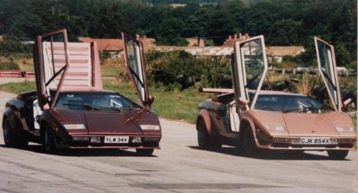 Countach  - Page 9 - Lamborghini Classics - PistonHeads - In the image, two Lamborghini sports cars are parked side-by-side on a gray paved road. The car on the left is a vibrant brown metallic finish, while the car on the right has a sleek copper color. They both feature the iconic design of gull-wing doors, which are open in this scene. The background shows a lush, grassy field and trees, suggesting a rural or semi-rural setting. The cars are on a quiet road, with no other vehicles in sight, indicating a peaceful and serene environment for the high-performance vehicles.