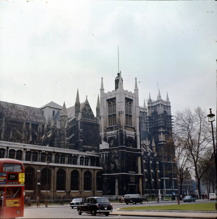 A 'period' classics pictures thread (Mk II) - Page 137 - Classic Cars and Yesterday's Heroes - PistonHeads - The image captures a scene in London, England. Dominating the frame is a majestic Gothic cathedral with two towers and several spires reaching towards the sky. The architecture of the building is intricate and ornate, indicative of its historical significance. In the foreground, there's a bustling street filled with traffic, including cars and a double-decker bus. The presence of a red telephone box adds a touch of modernity to the otherwise historic scene. Despite the urban setting, the cathedral stands as a testament to London's rich history.