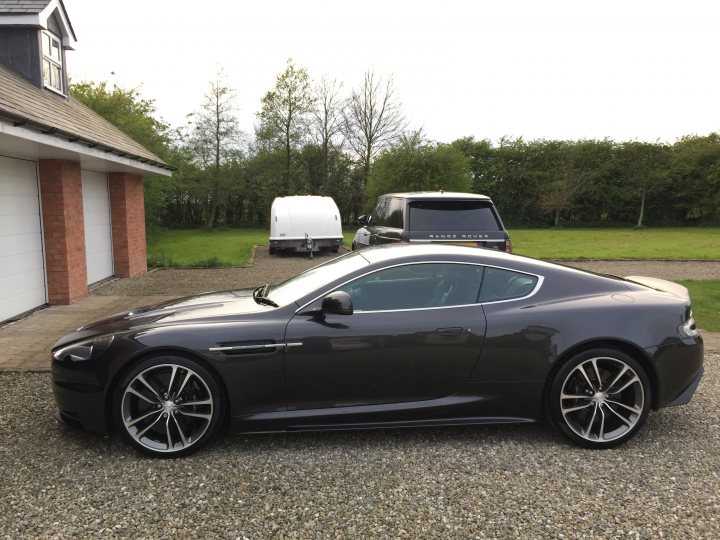 Our other cars - Page 5 - Aston Martin - PistonHeads - The image showcases a sleek, black sports car parked on a gravel driveway. The car is positioned facing towards the right side of the frame, with a side view that accentuates its curved design and black wheels. A garage is visible in the background, indicating that the location is likely residential. In the background, there's another vehicle and a utility vehicle, suggesting the presence of other property uses. The weather appears to be overcast, with a hazy, gray sky overhead.