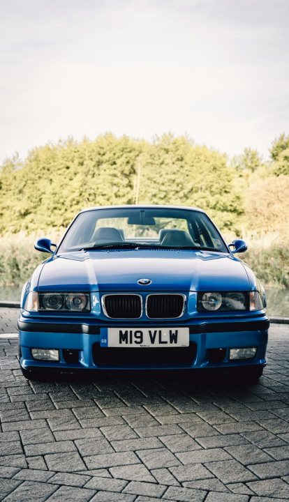 BMW E36 M3 - Reckless Restoration  - Page 10 - Readers' Cars - PistonHeads UK - The image is a split-screen photograph. On the left, there's an unattended blue BMW car parked on a brick pavement with grass and trees in the background. The car has a distinctive white stripe running down its side. On the right, there's a blurred view of a person taking the photo from a low angle, partially obscured by the car's shadow.