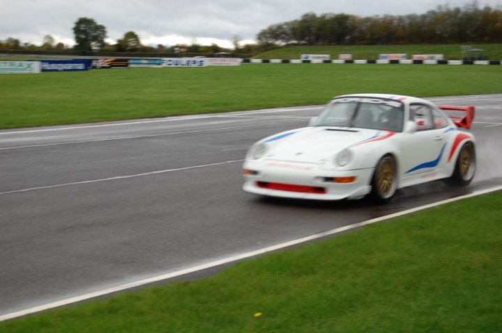 Pistonheads - The image captures a moment of exhilaration on a track. At the center of the frame is a white Porsche race car speeding along the wet asphalt. The car's sharp handling is evident as it takes a sharp right turn, the tires touching the grass outside the track. The perspective of the photo suggests it was taken from a spectator position, giving a sense of the thrill of the race. Despite the overcast sky overhead, the scene is full of energy and motion.