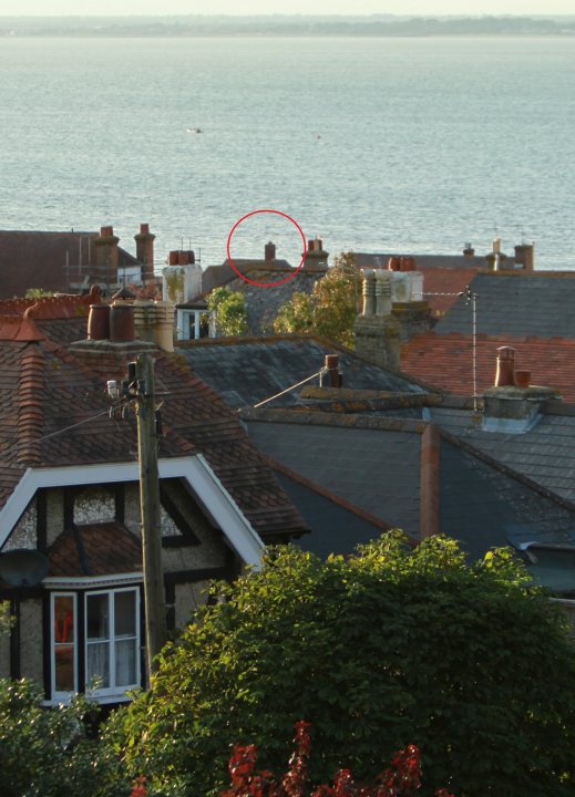 A large building with a clock tower on top of it - Pistonheads - The image shows a residential area featuring prominently shingled rooftops, either in warm tones or black. In the red-toned rooftop circle area, there appears to be a chimney with round accessories on the rooftop of the house. The area is elevated and provides a clear view of a body of water below the houses, suggesting a coastal or waterfront community. The sky is blue, suggesting that the photo might have been taken during a clear day.