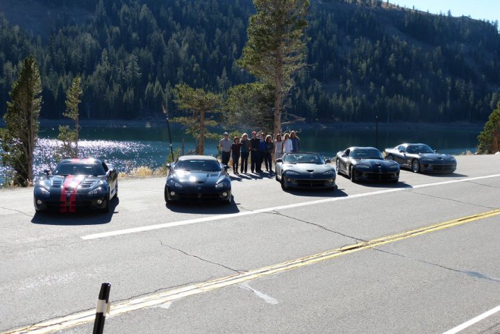 A group of cars driving down a street - Pistonheads - The image depicts a group of people gathered on the side of a road, with multiple sport cars parked in a line along the road between them. The setting appears to be outdoors, near a lake or waterway, with a scenic backdrop of forests. The day appears to be sunny, as light is visibly shining on the surface of the water and the cars' exteriors. The cars are sleek, and they stretch from the foreground of the image to the background, as if forming a series that blends both color and perspective.