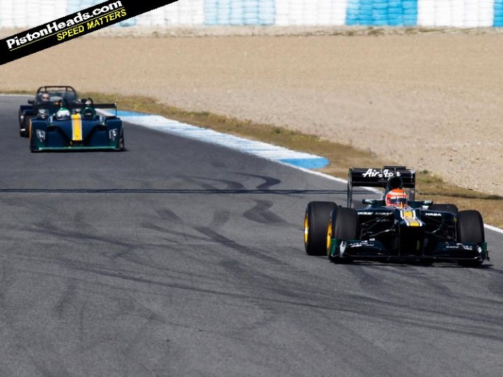 My Caterham R500 with the Caterham F1 team in Jerez - Page 1 - Readers' Cars - PistonHeads - The image captures a thrilling moment of two race cars speeding around a race track curve. The cars, each with a driver, are leaning into the turn, a testament to the high speeds they are reaching. One car slightly leads over the other, both adorned with partial sponsor logos. The setting appears to be a dry day on a circuit with a long straight stretch followed by a left-hand turn. The presence of the watermark suggests the image is from a real-life racing event report.