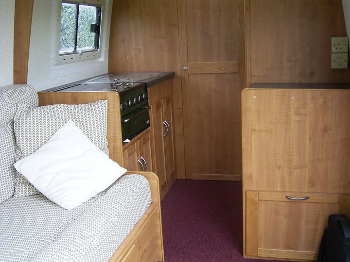 Camper Pistonheads Talk Vans - The image depicts the interior of a small living area with a cozy atmosphere. Two wooden chairs with comfortable pillow cushions are facing each other, inviting conversation. A red carpet adds a pop of color to the warm tones of the wood. A cabinet next to one of the chairs includes a sink, suggesting a combined living and kitchen space. A window with white trim lets in natural light, giving the space a relaxed and welcoming feel.