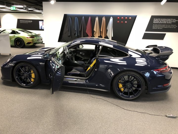 991.2 GT3 - Colours. Spec. Q+A. etc etc.. - Page 296 - 911/Carrera GT - PistonHeads - The image depicts a scene from a car dealership, showcasing a new Porsche Cayman sports car. The car is a dark blue with yellow accents, parked on a black carpet that contrasts the vehicle's color. The dealership is adorned with showroom decor, including a display of ties in the background. The precise positioning of the car, highlighted by a red carpet leading to it, suggests it's the centerpiece of the showroom. Additional information about the car, such as its model, features, and pricing, is not visible in the image.