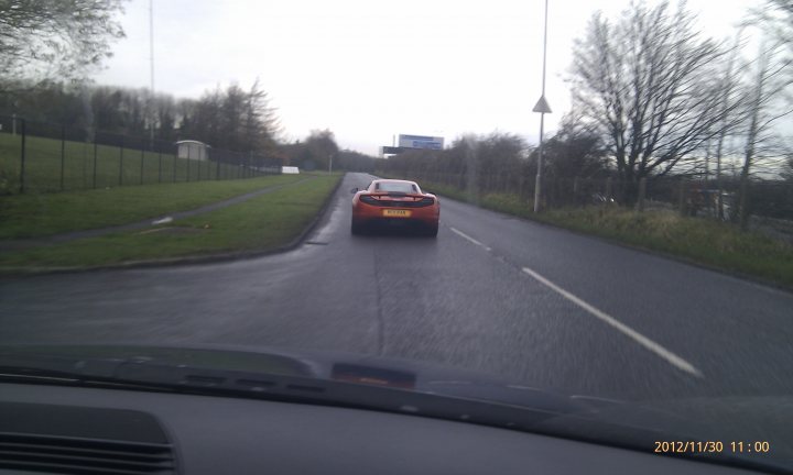 McLaren Spotted Mallusk 30th Nov - Page 1 - Northern Ireland - PistonHeads - The image depicts a single-lane road where a red car is driving away from the viewer. The road is wet, implying it may have rained recently. To the right of the image, there is some greenery, suggesting a rural or suburban setting. In the distanced background, there appears to be a sign or billboard, although the text is not legible. The date and time stamp on the image read "2012/NOV/30 11:00," indicating that the photo was taken at 11:00PM on the 30th of November, 2012.