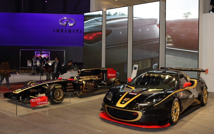 Looking for Lotus Evora stripe/wheels gold colour code - Page 1 - General Lotus Stuff - PistonHeads - The image showcases an exhibition floor with a prominent display of two luxury cars, one with a metallic and the other with a gold and black color scheme. Both cars are high-end models adorned with the logo "INFINITY." A larger-than-life reflection of a car can be seen on glass near the vehicles, contributing to the grandeur of the display. There's a white wall in the background where the INFINITY brand is prominently displayed. A handful of individuals are scattered around the room, some standing close to the cars, possibly observing or discussing the vehicles.