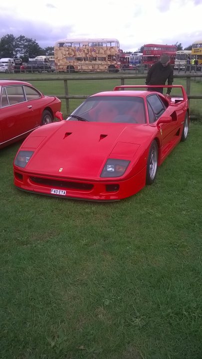 Classic Ferrari at Croft today. - Page 1 - Ferrari Classics - PistonHeads - The image showcases a vibrant red Ferrari sports car prominently parked in a lush green field with a wooden fence enclosing it. The vehicle, characterized by its wide body, sharp angles, and the iconic prancing horse emblem, is facing towards the right side of the photo. Numerous other cars, including another Ferrari and a bus, are visible in the background, suggesting a potential event or gathering. Additionally, there are a few people present in the distance, adding a sense of scale to the scene. The sky above is partly cloudy, hinting at an overcast or possibly changing weather conditions.