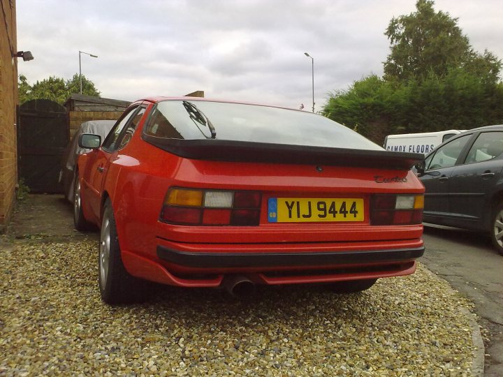 Didnt Exist Mxs Pistonheads - The image shows an older red German car parked on a gravel surface in front of a building. The car has a yellow license plate that reads "YIJ 944". It appears to be a two-door model, likely a coupe or sportscar, given its sleek and aerodynamic design. On the curb next to the car, there's another vehicle partially visible. The sky is overcast, suggesting it may have been a cool or cloudy day when the picture was taken. The view is from ground level at the back end of the red car, focusing on the back bumper and rear lights.