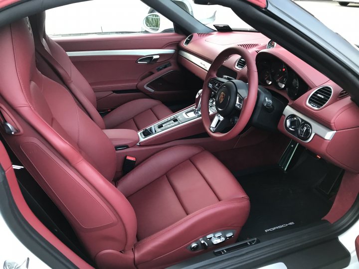 The image shows the interior of a sports car, viewed from the back seat. The seats are adorned with red leather upholstery and have cup holders on the armrests. The steering wheel is visible in front of the passenger seats, featuring an infotainment screen at its center. The dashboard and door panels are also visible, exhibiting a combination of materials including wood and metal finishes. The car's interior design suggests luxury and comfort.