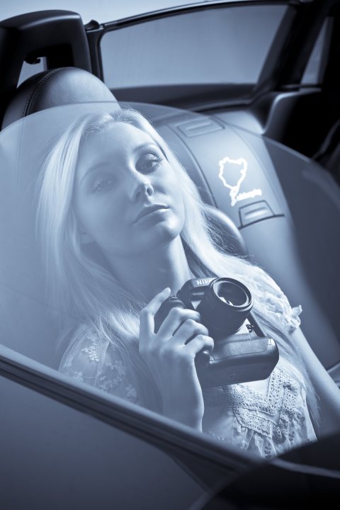 Pistonheads - This is a photograph featuring a young woman reclined in a car seat, looking out a car window with a relaxed and thoughtful expression. She possesses light-colored hair and is wearing makeup. Her hand is on her chin in a contemplative position. There is a smartphone displayed near her, reflecting on the car seat's surface, suggesting she might have taken a selfie. The car's speedometer is visible on the dashboard. The image style appears to be a blend of realism with a slightly desaturated color palette, adding a cinematic effect.