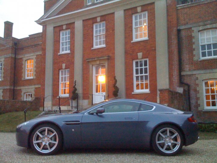 Testing the Water - MY07 Vantage - Page 1 - Aston Martin - PistonHeads - The image features a dark gray Aston Martin sports car parked in front of a large, old-style building. The building has a rustic charm, with its noticeable brickwork and multiple windows. The setting appears to be a residential area with well-maintained yards and a well-paved driveway. The car is positioned next to a pillar, illuminated partially by sunlight, suggesting it is either morning or mid-afternoon. The vehicle's reflective surfaces mirror the building and the ambient light, adding to its sleek and dynamic appearance.