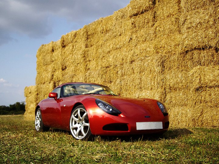 Pistonheads Colours Interesting - The image showcases a vibrant red sports car parked in front of a wall of golden hay bales. The car, a convertible with the top down, is situated in what appears to be a field with tall grass, enhancing the rural setting. The sky in the background is overcast, casting a soft light over the scene. The contrast between the sleek, modern car and the traditional hay bales creates an interesting juxtaposition.