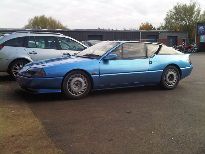 Picked up this on Friday - Page 1 - French Bred - PistonHeads - The image features a blue convertible parked in a gravel parking lot, with another car and some buildings in the background. The sky above is overcast. The car appears to be of a classic or vintage design, possibly from the 1990s. The vehicle's bodywork reflects the light slightly, highlighting the blue color and the car's journey, as indicated by small rocks and debris underneath the tires. The overall setting suggests a calm day in an urban or suburban environment.