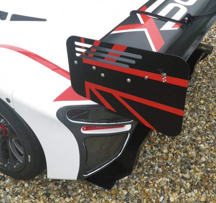 What's going on with bulkheads then? - Page 1 - Radical - PistonHeads - The image shows the rear end of a car with distinctive aerodynamic designs. Dominating the lower half of the frame is a robust, rear spoiler with a prominent design featuring a red and black striped pattern, possibly a decal or adhesive film. This design structure is often seen on vehicles designed for racing or high-performance driving. Below the spoiler, partially visible, is a vent or cooling grille with a mesh pattern, which is likely part of the aerodynamic package for enhancing overall performance. The car body also features the Red and X logo, which is characteristic of some high-performance models, suggesting this is either an official team car or a heavily modified vehicle for racing or aesthetic purposes. Finally, the tires appear to be performance oriented, with a wider profile suggesting a higher grip capability. There's no visible text or branding on the car in this image.