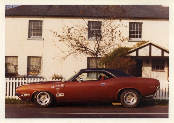 Dodge Challenger vs Charger - Page 1 - Yank Motors - PistonHeads - The image is a graphic photograph taken in front of a house with a two-car garage and a white picket fence. The main subject is an older-model red firebird coupe, likely from the 1960s to 1970s, parked on a street in front of the house. The car has a black roof, and there are various personal or identification stickers on the trunk and back window, giving the image a nostalgic or personal feel. The house is a two-story structure with a pitched roof and double-hung windows visible. The overall style of the image suggests it might be a vintage or personal photograph.