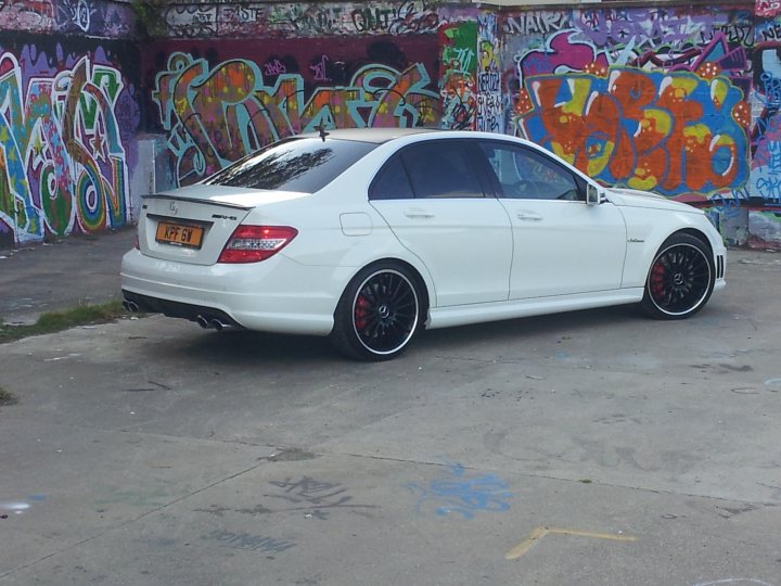 A white car parked in front of a building - Pistonheads - The image captures a vibrant street scene featuring a white Mercedes Benz, painted in ornate purplish designs, parked next to a concrete sidewalk. The sidewalk showcases a stylish backdrop of graffiti art, adding an urban touch to the tableau. The vehicle is adorned with a black body kit and sleek black wheels, enhancing its sporty appearance. The sky above is clear and blue, suggesting a bright and sunny day, further illuminating the vivid colors and design elements of the car and its surroundings.