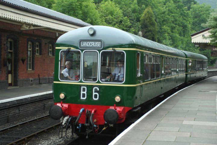 Pistonheads - The image shows a vintage-style train parked on tracks next to a platform. It's a green and cream train car with the number 6 on the front, indicating it might be part of a series or a specific model. The platform is adjacent to the tracks, suggesting this is a station where passengers can board and alight from the train. There are some people present near the platform, possibly waiting for the train or just passing by. In the background, there's an indication of a natural landscape with trees visible, giving the scene a serene atmosphere. The setting appears to be in daylight, as suggested by the lighting and shadows cast.