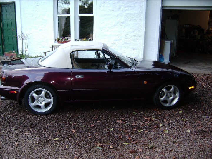 My Vr ltd comb A arrived today  - Page 1 - Mazda MX5/Eunos/Miata - PistonHeads - The image displays a burgundy-colored sports car parked on a gravel driveway in front of a white two-story house with green window frames. The car appears to be a classic model with a soft top, which is down and tied back. The photograph shows that the car is in good condition and presents a clear view of its side profile and part of the front end. The house in the background exudes a homey vibe, and the gravel driveway adds to the overall charm of the scene. The setting implies a quiet residential area.