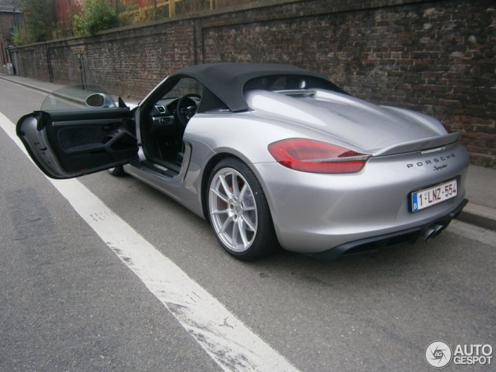 A car is parked on the side of the road - The image showcases a silver Porsche sitting by the sidewalk, displaying a striking personality with its black soft-top roof. The vehicle features a license plate that reads "1. LN-853", hinting at its origin. The Porsche is positioned on the right side of the street, adjacent to a brick wall, adding an urban touch to the scene.