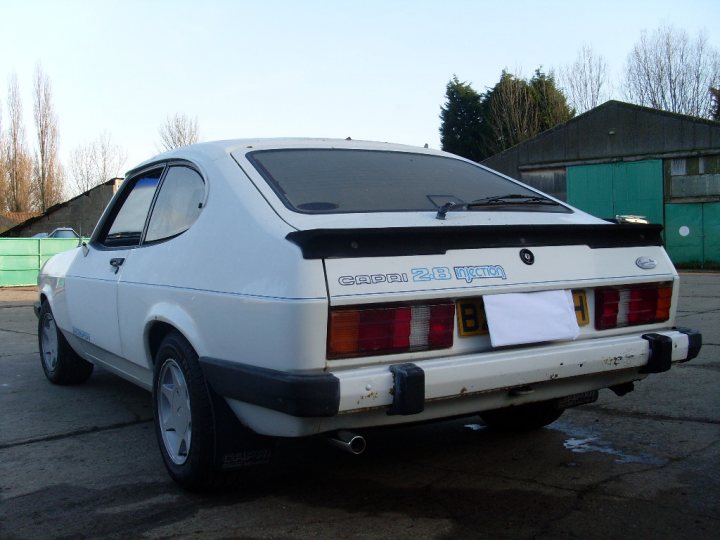 Starter Classic Pistonheads - The image shows a white hatchback car parked atop a paved area, possibly a parking lot. The car has a visible license plate with blue letters and numbers, as well as a spare tire affixed to the rear bumper. The body of the car exhibits signs of wear or recently repaired sections, indicated by the combination of new and old paint. In the background, there are buildings with green and grey walls, suggesting this parking spot is located near some sort of facility or establishment. The sky is largely overcast, and no people are visible in the immediate area of the car.