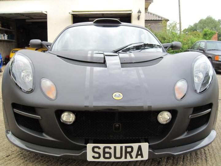 Plate Pistonheads Ive - The image shows a sleek, black sports car parked outdoors in front of a building. The vehicle has a smooth, modern design with a clear windshield and circular headlights. There's a front license plate that reads "S6USRA," and the grille features a badge with a 'Porshe' emblem, indicating the make of the car. In the background, you can see some benches and other vehicles, suggesting that this might be a public parking area or a showroom.