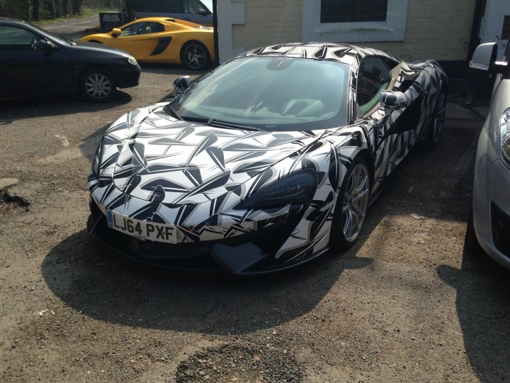 McLaren 570S - Page 6 - McLaren - PistonHeads - The image shows a sports car, which appears silvery and decorated with a black and white pattern. The car is parked on a gravel lot with a faded asphalt around the concrete. In the background, there are two other cars, one of which is yellow, parallel parked. The setting suggests an unoccupied driveway or storage area. The positioning of the cars indicates they are not currently in use.