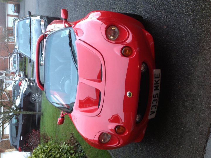 My first Elise S1 - Calypso Red - Page 1 - Elise/Exige/Europa/340R - PistonHeads - The image depicts a vibrant red sports car parked on a road. The car is positioned behind another vehicle, giving a sense of depth. The license plate on the red sports car is clearly visible, with "33" likely being part of a registration number along with "MKE". The vehicle is capturing the viewer's attention, possibly due to its shiny surface reflecting the surrounding environment. The car's design, presumably a German sports car model, is prominent with its distinctive shape and sleek lines.