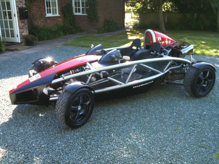 In the club - Page 1 - Ariel - PistonHeads - The image shows a red and black three-wheeled motorcycle parked on a gravel driveway. It features two wheels at the front and one at the back, which is covered but explicitly labeled as such. The motorcycle has a triangular structure with a seat that can accommodate at least two people. The background includes a house with a visible doorway and greenery, indicating a suburban setting. The sunlight is evidently cast on the gravel, suggesting it is daytime.