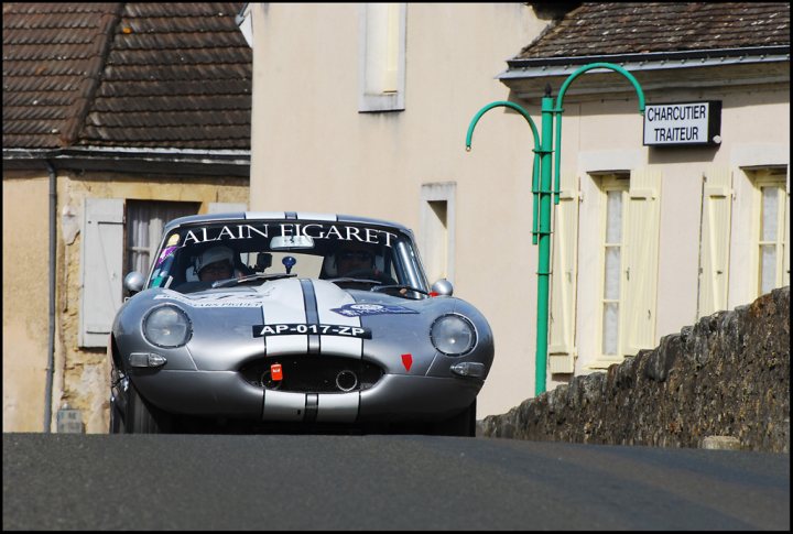 The Tour Auto came to town - Page 1 - Classic Cars and Yesterday's Heroes - PistonHeads - The image features an old racing car traveling down a curved street. The car is sleek and convertible, painted in shades of grey and white with a black roof. On its side, it's emblazoned with blue and white text, although the specifics are not fully legible. The body of the car shows signs of use and speed, with faded paint and visible screws or rivets. In the background, there is a similar, smaller vehicle parked to the side of the street. The street itself has a cobblestone-like surface, adding a charming rural feel to the scene.