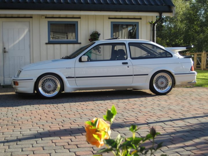 So what car have you been obsessing about today?? - Page 115 - General Gassing - PistonHeads - The image features a sleek, white sports car parked on a brick-paved driveway of a white house. The car has a prominent air intake at the rear, obscuring a part of the tailgate that would typically be the trunk. In the foreground, there is a small orange flower and a few plants or bushes to the left side of the image. The overall scene suggests a residential area and might imply the car is either being used for leisure or is parked there for a short duration.