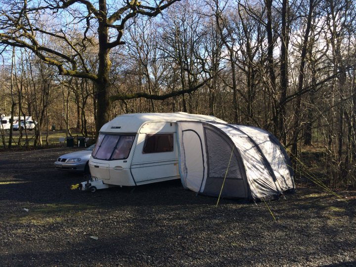 Awning Problems - Page 1 - Tents, Caravans & Motorhomes - PistonHeads - The image showcases a tranquil outdoor setting with a campsite, a tree, and two cars. Dominating the scene is a white, unoccupied caravan, prominently positioned in the foreground with its front door open, giving the impression of recent use or empty preparation for another journey. 

The caravan is pitched on a gravel surface, ready for exploration, though no tents are currently set up. In the background, there's a solitary tree providing a sense of natural tranquility to the scene, juxtaposed with the man-made caravans and cars. 

The sky above is a clear blue, suggesting a bright and sunny day, perfect for an adventure or a road trip. Adding to the sense of adventure, there are two vehicles parked in the background, one of them being a car. The second vehicle is an RV, larger than the truck and car in the background, adding variety and depth to the image. Overall, the image captures a moment of pause before or after a camping trip.