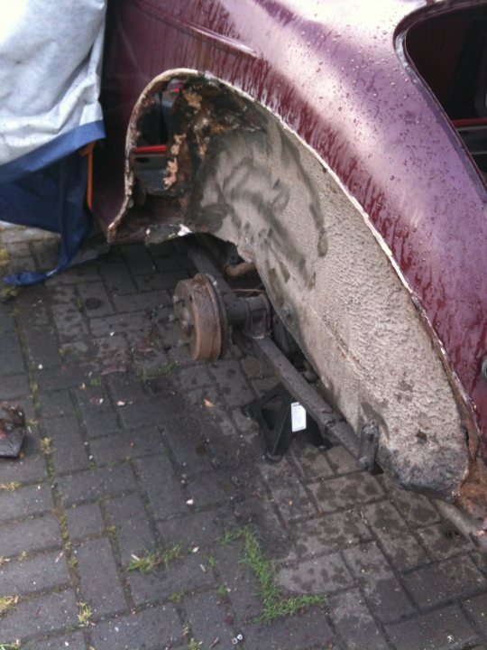 1971 Morris Minor 1000 Resto thread (Maybe) - Page 1 - Classic Cars and Yesterday's Heroes - PistonHeads - The image shows a close-up of the wheel and suspension area of a dark red car's right rear corner. There is visible damage: the wheel hub is damaged with burnt paint, the tire rim is bent and scratched, there are bruises on the bodywork, and the wheel is dislodged, revealing damaged parts. The ground is paved, and there are sparse green plants growing through some cracks. This is a snapshot of the aftermath of a car accident.