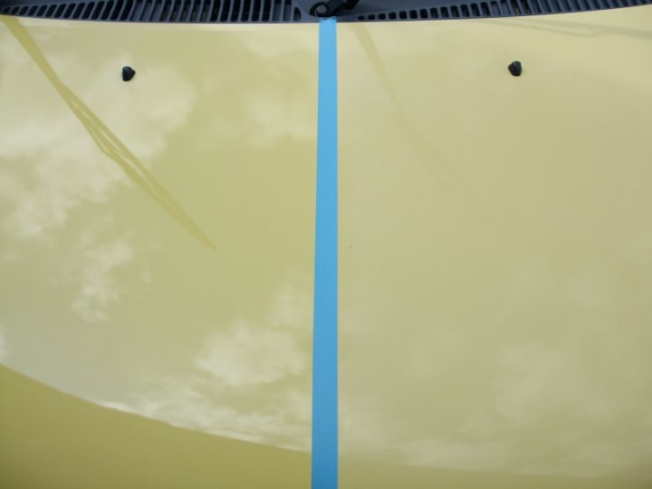 Shiney Pistonheads - The image shows a close-up view of a car, focusing on its front fender and hood. The fender of the car is painted a bright yellow, and the hood has a reflection of a cloudy sky. A prominent, dark-colored object is located in the middle of the fender, positioned exactly between two black circles. On the right side of the fender, a horizontal, blue line runs parallel to the smooth, reflective surface of the car.