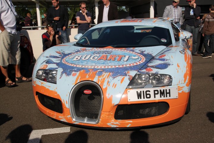 Supercar Sunday pictures - Page 2 - Goodwood Events - PistonHeads - In the image, a vibrant Bugatti coupe is the focal point. The car is not an ordinary one; it has an elaborate and artistic body paint in shades of blue and orange. The Bugatti, with its unique design, appears to be a part of a sprint or race event as suggested by the colors and possibly a decal on the side that reads "Bugatti". The car is surrounded by several individuals who are standing at a safe distance. It appears to be parked in a spacious parking lot, and the scene seems to be outdoors during a sunny day.