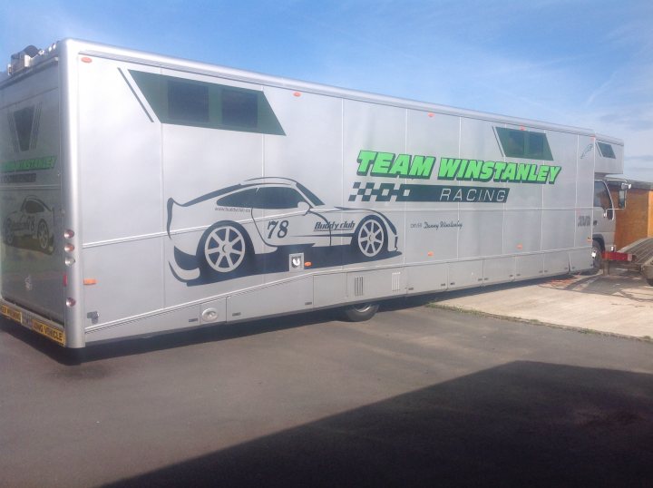 race transporter advice - Page 1 - UK Club Motorsport - PistonHeads - The image shows a large hauling trailer parked on an asphalt surface during daytime. The trailer is predominantly silver with a graphic of a car painted on its side. The artwork features a number '78' on the vehicle's door, and the text "TEAM WINSTANLE RACING" is visible, suggesting that it is associated with a racing team, perhaps in a motorsport context. The background is out of focus, but it appears to be a clear day with a few clouds, as no landmarks are distinctly identifiable. The style of the image is that of a real-world photograph, likely taken to document the trailer in its current location or context.