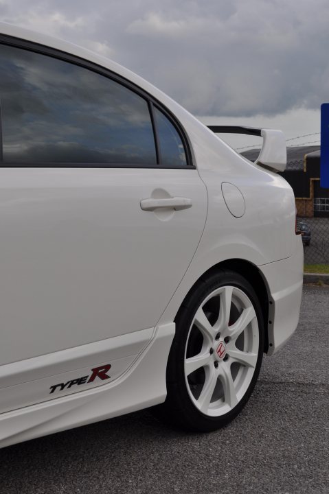 Pistonheads Type Import - The image features a close-up of the rear section of a silver or white sports car. The car's bodywork is noticeably modified with aerodynamic enhancements, including a protruding wing over the rear door and a spoiler above the trunk lid. The vehicle's left rear wheel is prominently visible, displaying the "TYPE R" logo on the center spokes. The ground appears to be asphalt, and the sky is overcast, suggesting the photo may have been taken in overcast weather.