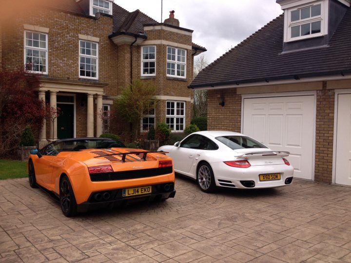 Performante Edizone Tecnica  - Page 1 - Gallardo/Huracan - PistonHeads - This image captures a serene residential scene with two cars parked side by side on a brick driveway. The driveway is lined with hedges and a tree, adding a touch of greenery to the scene. The house itself has a red brick facade, giving it a warm and inviting appearance. On the right side of the image, there's a light yellow car, and to its left, parked on the same driveway, is an orange convertible. Both cars are parked parallel to the house, which is framed by a collection of windows and a prominent white door. The overall scene is a beautiful representation of a peaceful residential area.