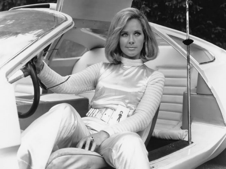 Pistonheads - The image is a black and white photograph featuring a woman sitting inside a car. She is positioned in the back seat and appears to be waiting or relaxing. The woman is wearing a sleek, shiny suit with a white top and black pants. In her hands, she is holding an umbrella. The car's interior is spacious, with visible luxury features such as leather seats and a raised back seat. The presence of a large umbrella suggests the car's design allows for unusual or accommodating passenger experiences.