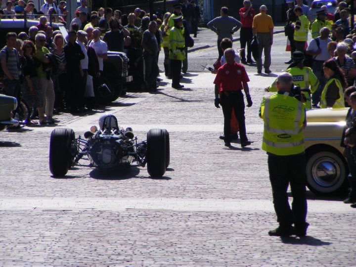 A group of people riding motorcycles down a street - Pistonheads - The image portrays a vibrant street scene where an old-fashioned car with large wheels is the center of attention. A crowd of spectators lines the sides of the cobblestone street, one of whom is taking a photograph. A race car is joining the parade, driving towards the crowd, and a police car is also present, escorting the vehicles on the street. The image captures the excitement and curiosity of the spectators and the uniqueness of the antique vehicle.