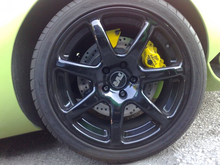 Options Exterior Pistonheads Colour - The image focuses on the wheel of a vehicle, showcasing its black rims with a metallic finish. Inside the wheel, a thick, yellow brake pad can be seen. The wheel is attached to a green tire and is positioned slightly out of view. The scene suggests a close-up of a wheel review or a detailed look at a vehicle's suspension system. The image captures both the aesthetic appeal of the rims and the functional parts of the vehicle's braking system.