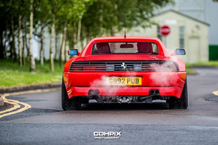 Ferrari 348 chit chat - Page 1 - Ferrari V8 - PistonHeads - In the image, a vibrant red Ferrari car is prominently featured, driving down a winding road. The car is coming around a corner, adding a sense of speed and dynamic movement. It leaves behind a trail of white smoke, adding an element of drama to the scene. The road itself appears to be well maintained, with clear visibility, suggesting a good day for a drive. In the background, a quaint white cottage with a clock on its side and trees lining the road contribute to the overall pastoral charm of the scene. The image is taken in such a way that it focuses solely on the car and road, providing a clear view and emphasizing its movements.